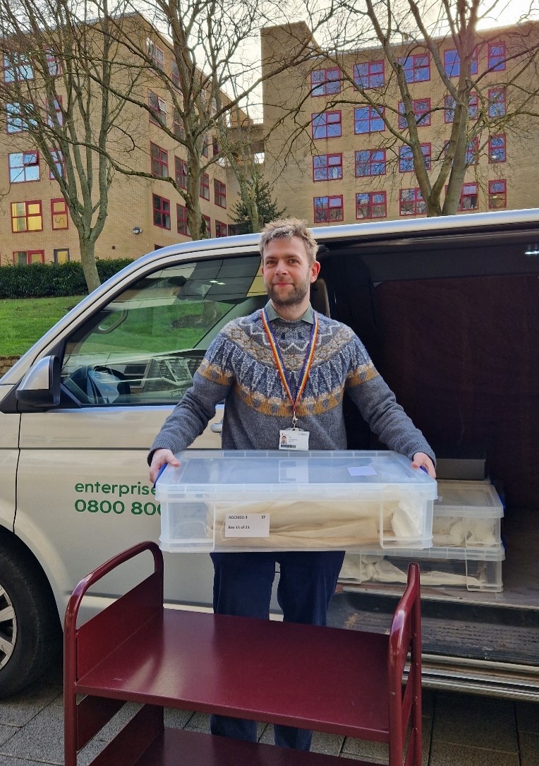 Photograph of man carrying a large plastic box of archive material, standing next to a trolley and a van containing similar boxes. University of Surrey buildings can be seen in the background.
