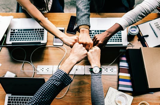 Five people's fists meeting each other over a desk of laptops and mobile phones