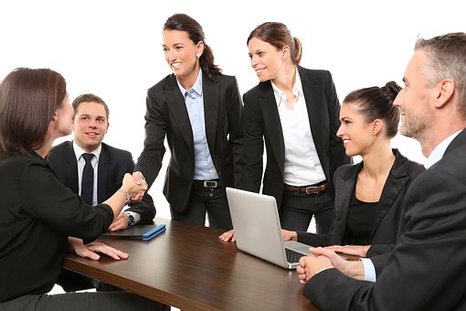 Six smiling people in suits (4 women, 2 men). Two of the women are shaking hands.