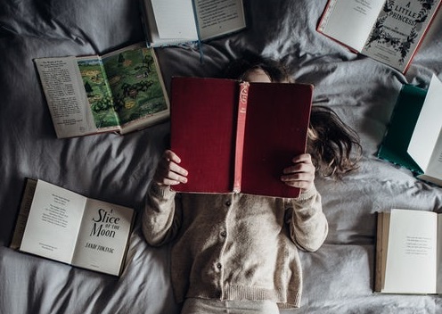 Girl reading large red book on bed surrounded by other books