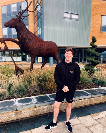 A photo of James standing by the Stag sculpture in the piazza outside the Rik Medlik building at the University of Surrey