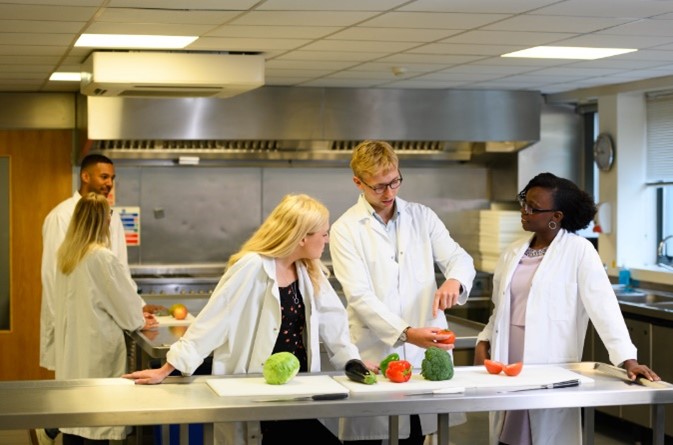 A group of workers wearing white coats at a bench, discussing the vegetables in front of them. Veg includes, broccoli, aubergine, cabbage and red pepper