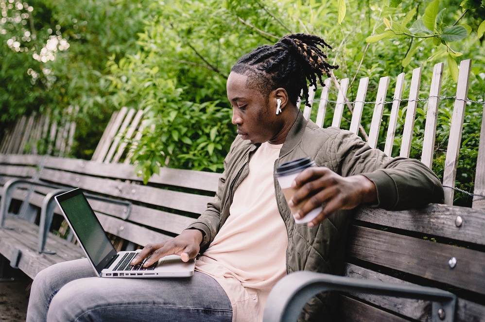 Placement student with dreadlocks sitting on a bench with a laptop and a coffee