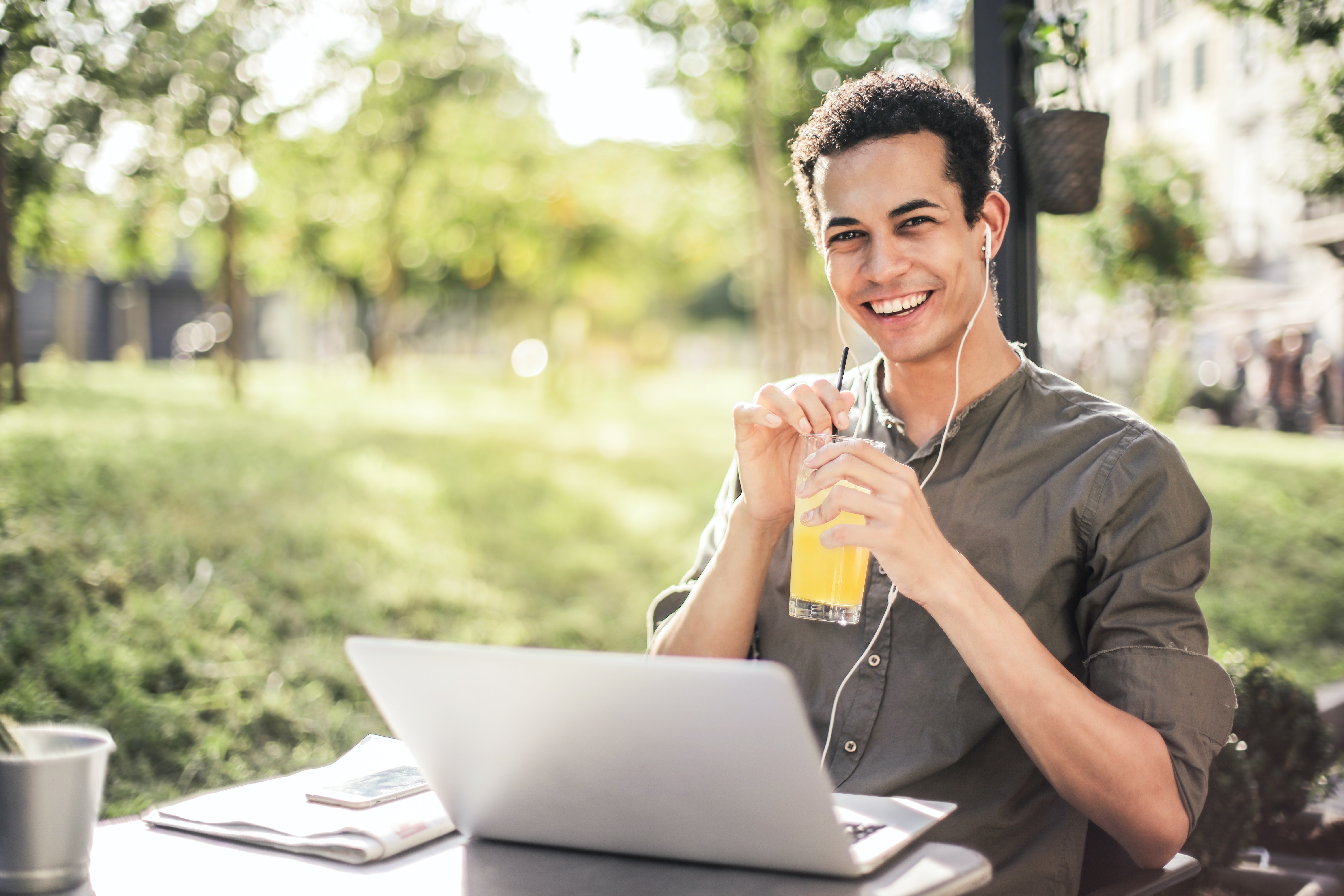 Young man sitting outside with a cold drink, he is on a laptop and has earphones in