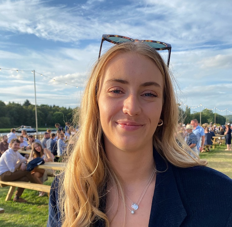 photo of Amelia on a sunny day. She has sunglasses perched on her head and there are lots of people sitting behind her. There is a river in the background.