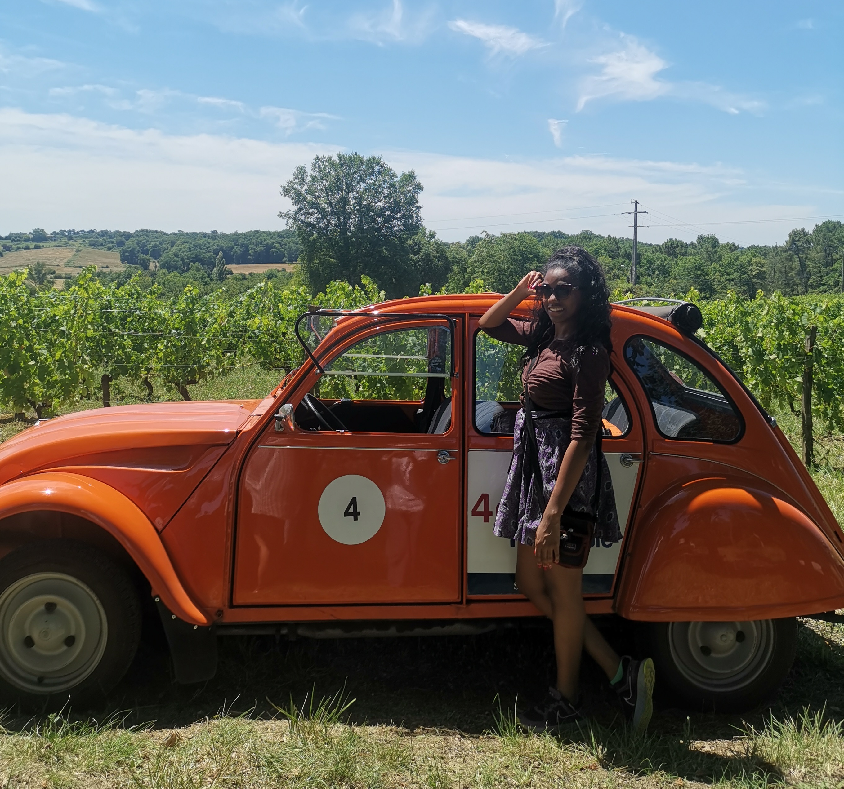 Rochelle wearing sunglasses on a sunny day, standing by a classic car in beautiful scenery near Paris