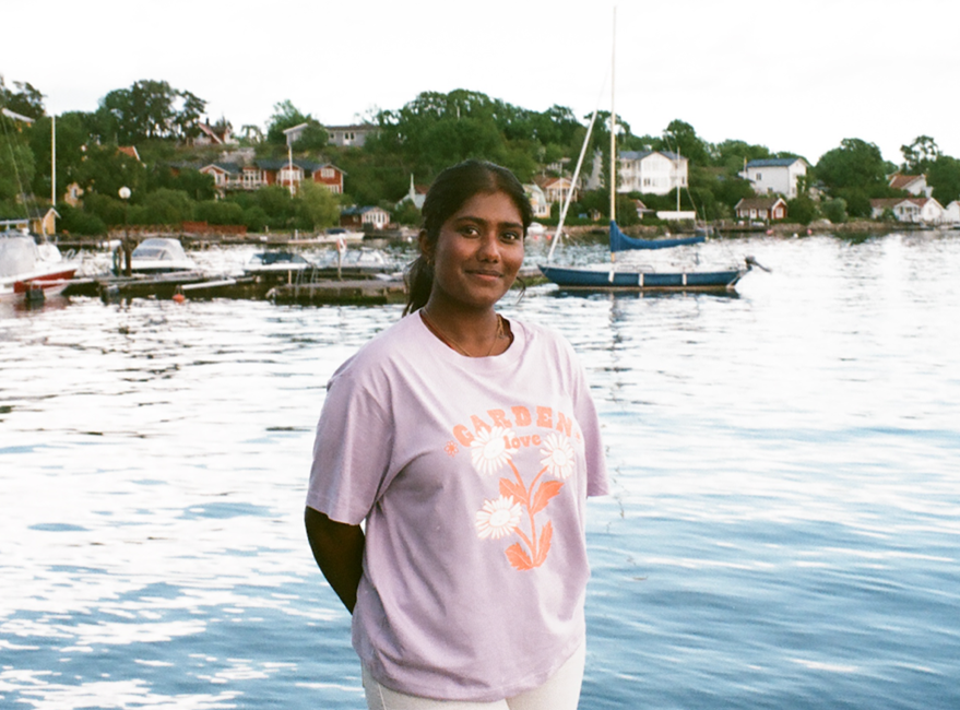 Sahini is smiling and standing in front of some water with boats on it, there are houses in the background