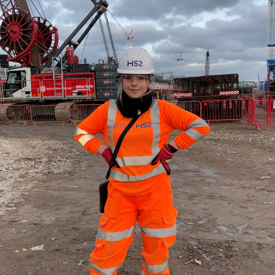 Hanna in high viz clothes and a HS2 hard hat in front of an engineering site.