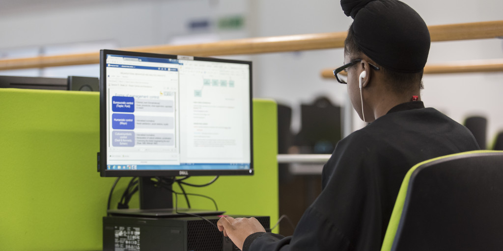 Photo of a student seated on a swivel chair at a computer with single screen situated on Level 1 of the University of Surrey Library. 