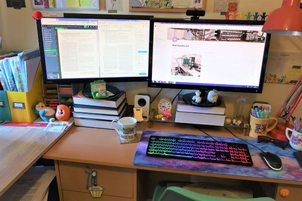 A desk with two monitors stnading on books. A colourful keyboard is sitting in front of the right hand monitor. There is a mug of tea next to the keyboard.