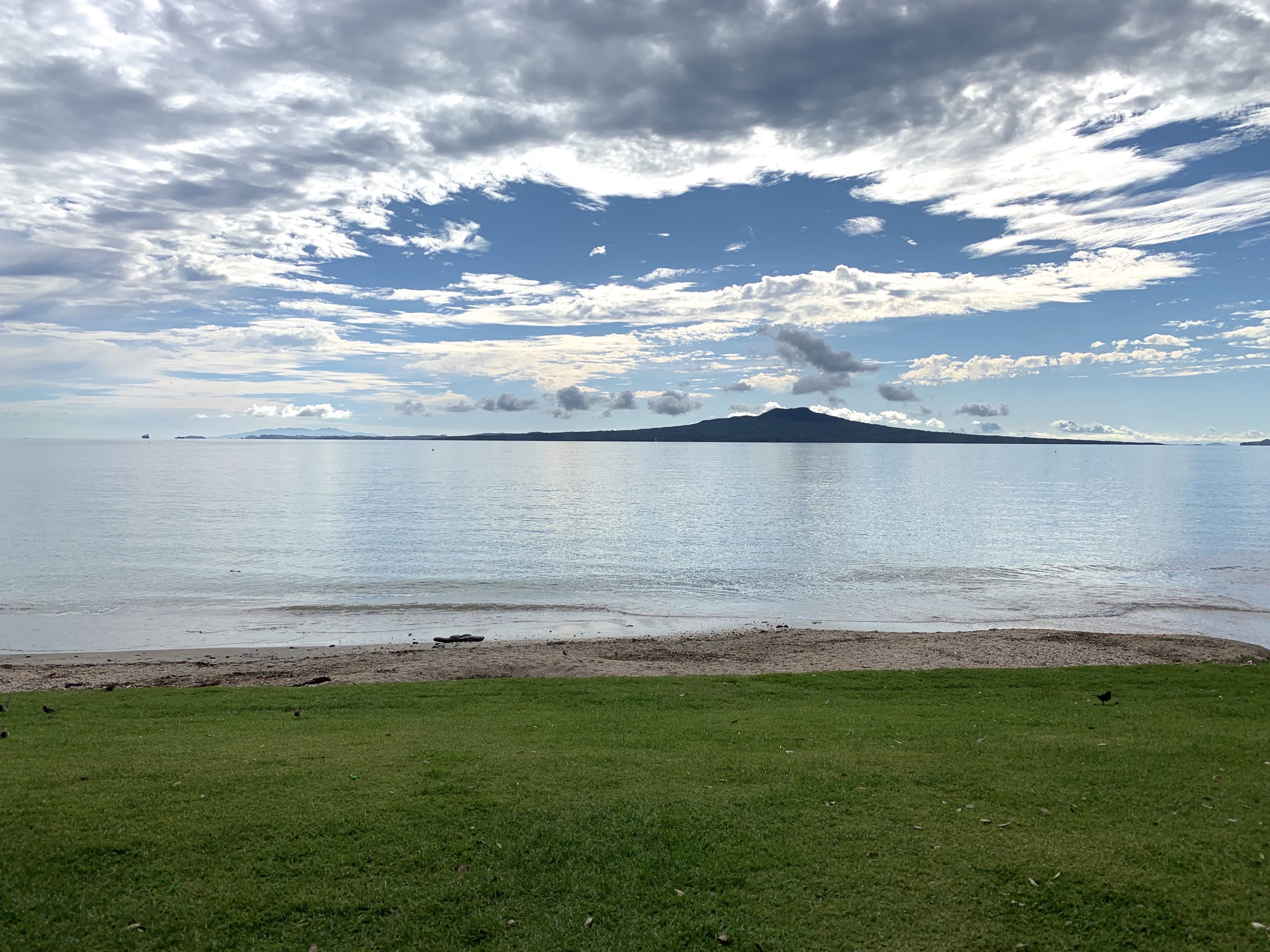 A beach with the tide in. Grass in the foreground and a volcano in the background.