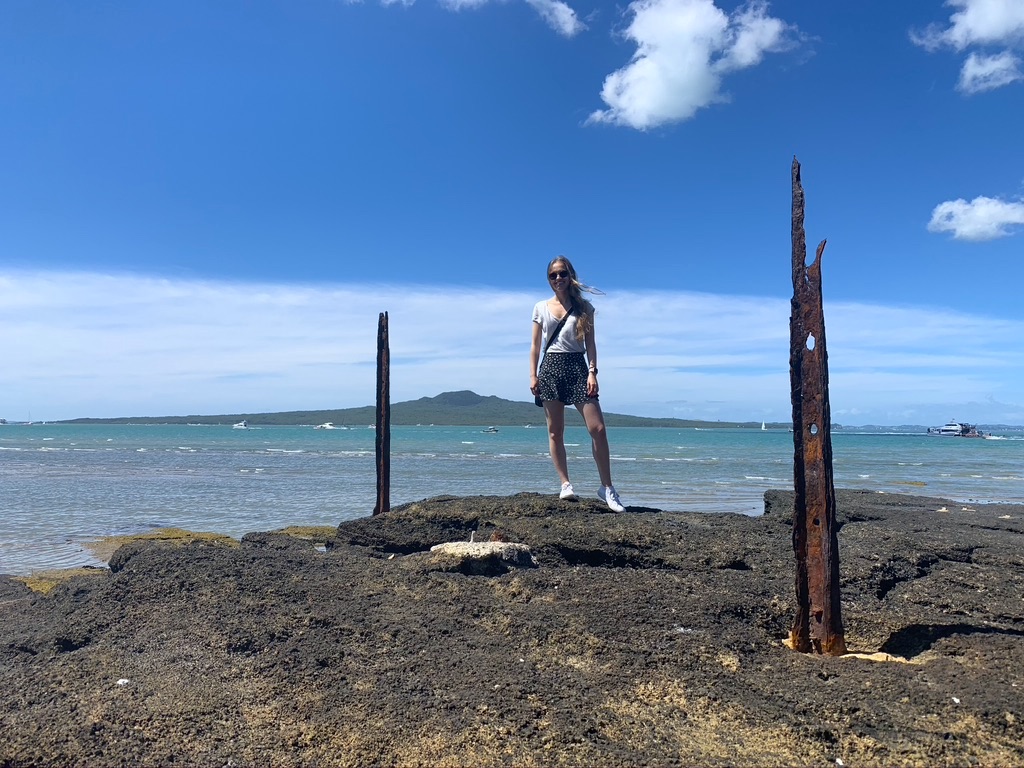 A girl standing on a rock, posing in front of the ocean and a volcano in the background.