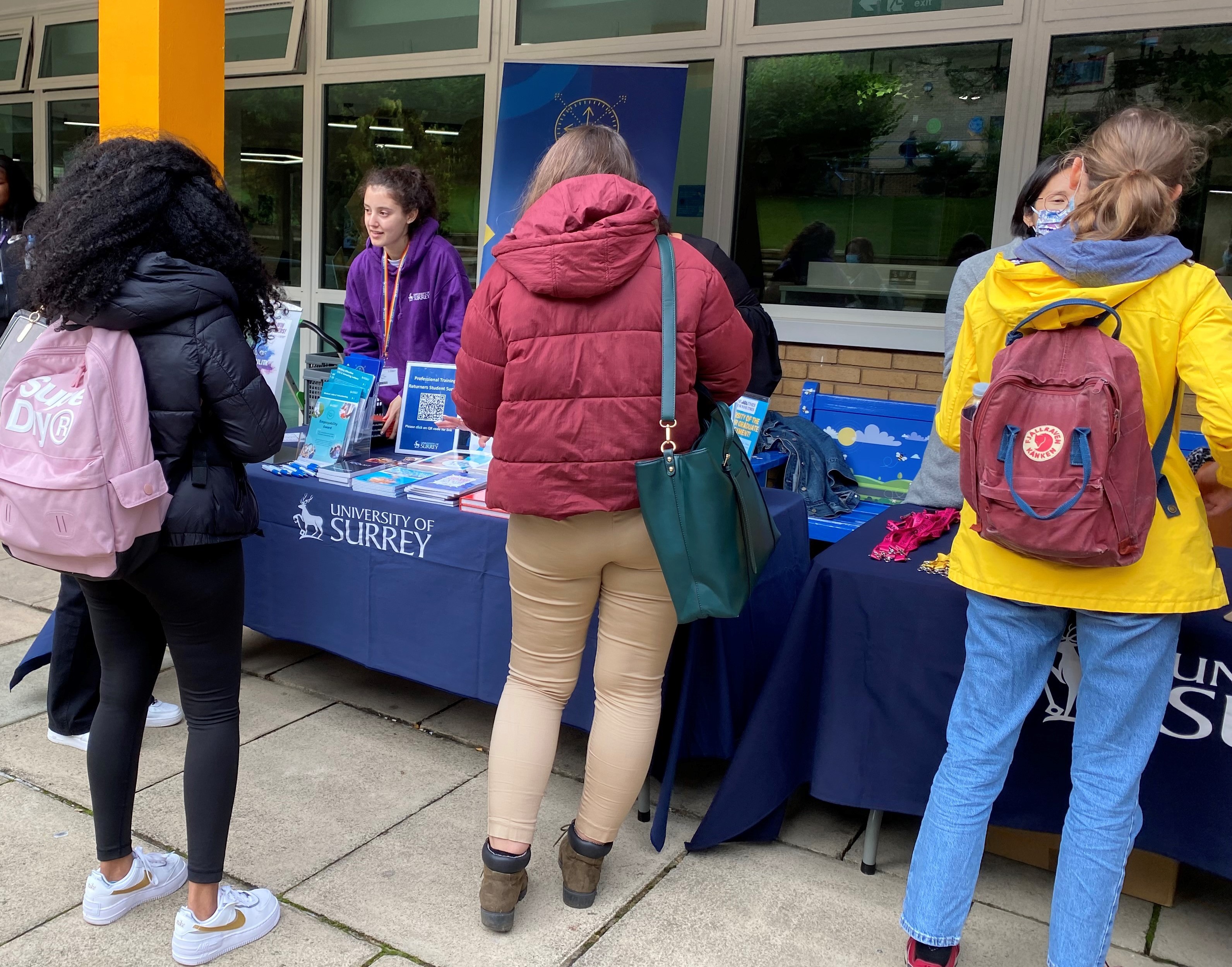 Image of Olive doing a pop-up stand outside the Hive. Several students in colourful coats are engaging in the stand.