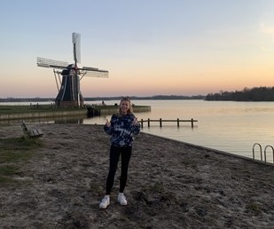Grace standing on a beach in front of a windmill in The Netherlands, the sun is setting behind the windmill