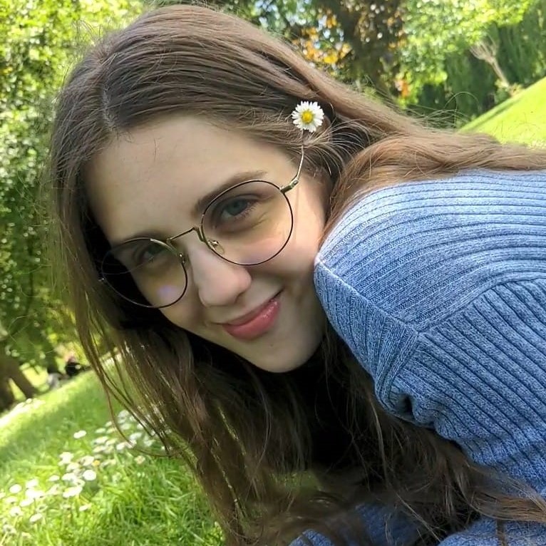 Close-up photo of Ivy sitting by the Lake. Ivy is a blond girl with glasses, and she is wearing a blue jumper
