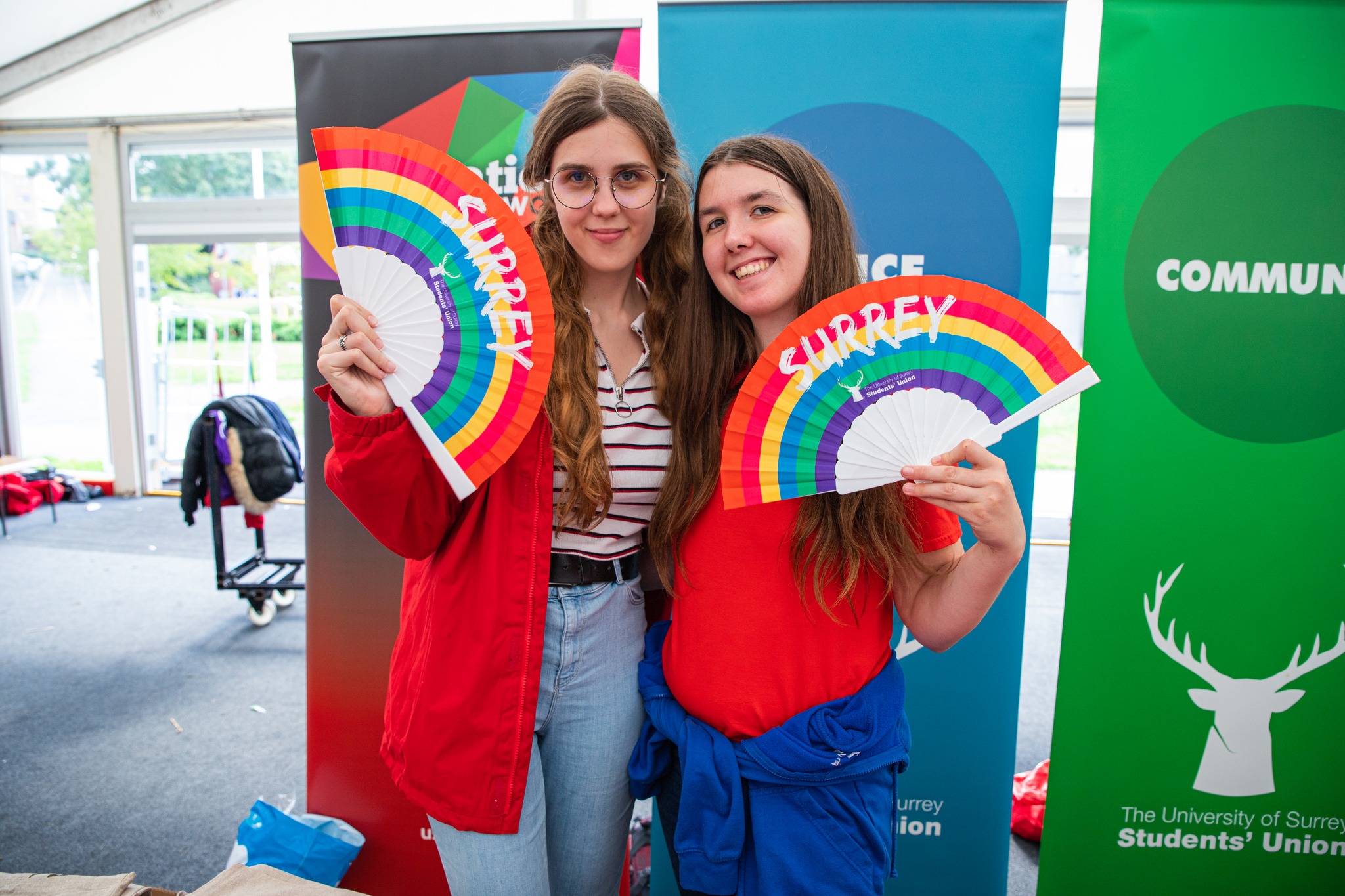 Two girls posing with rainbow-coloured fans in their hands. The girl on the left is tall and blond, with glasses, wearing white striped shirt and a red coat. The girl on the right is shorter with brown hair. She is wearing a red T-shirt.