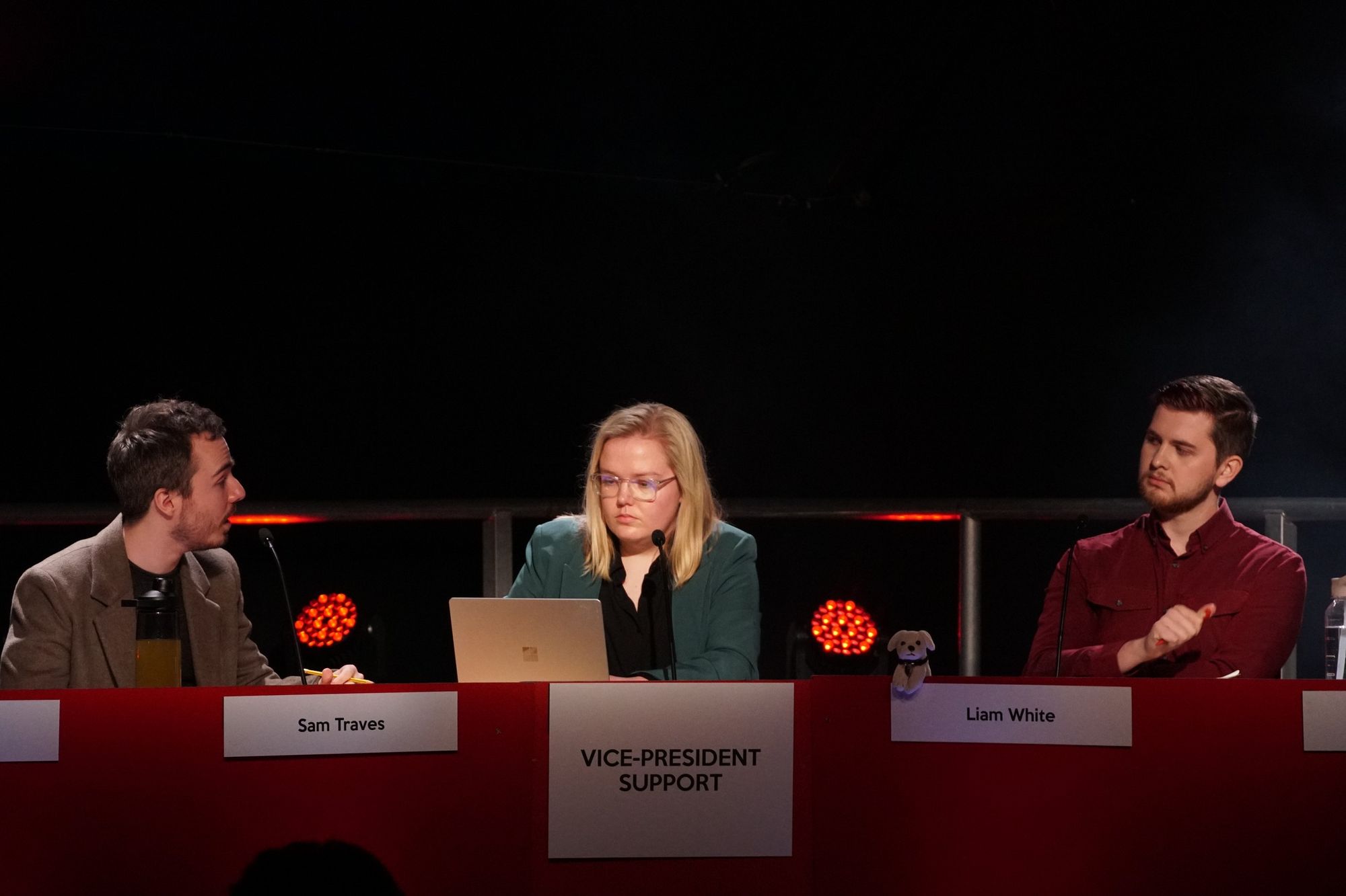 A young woman and two young many sitting on each side of her, on a stage, engaging in a debate