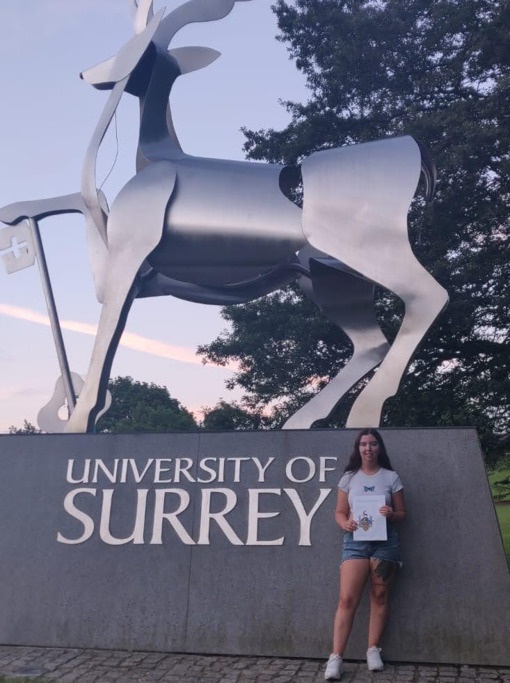 University of Surrey student smiling with dissertation research project in front of main university entrance with Stag statue.